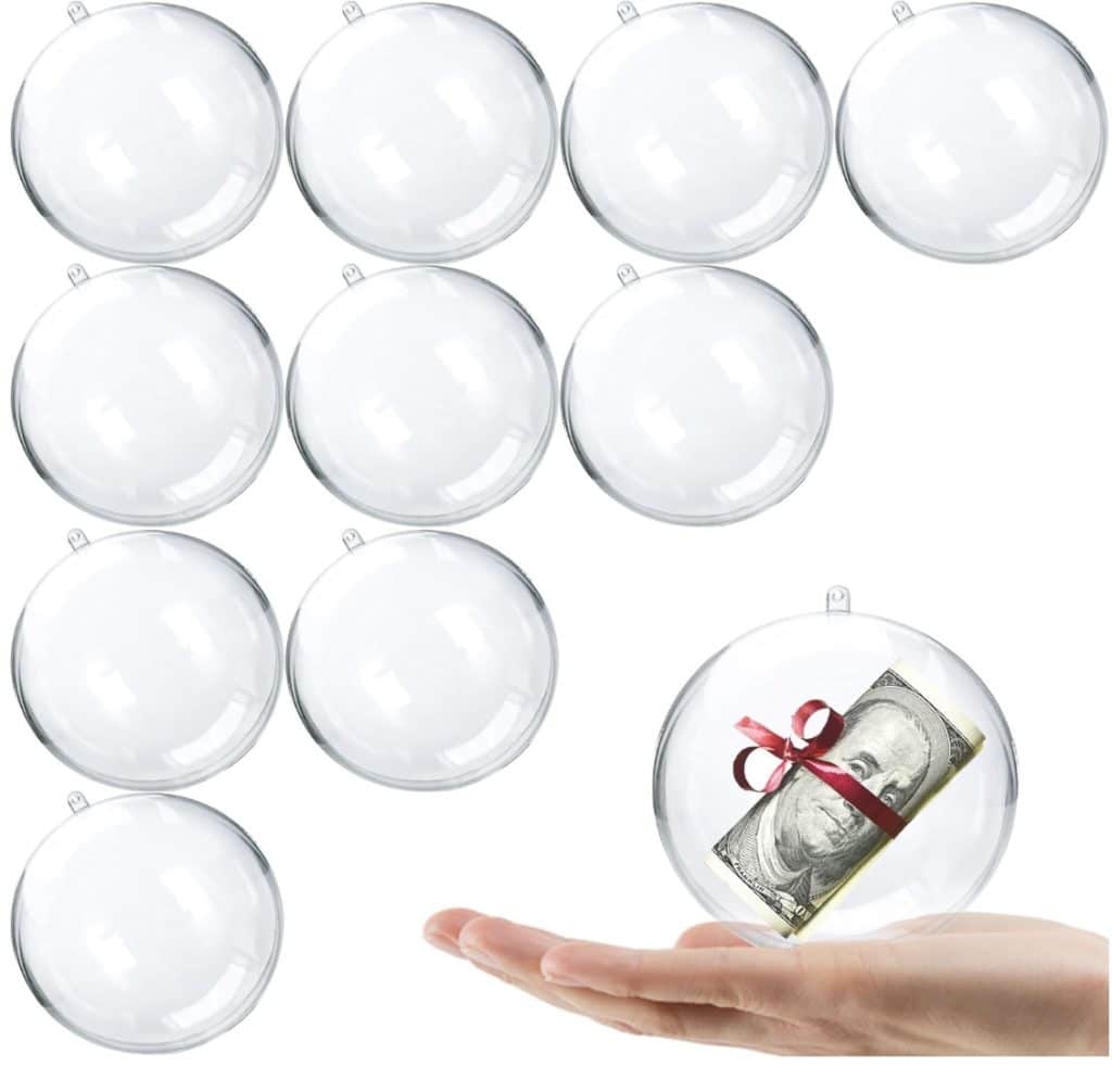 A hand holds a clear ornament with a rolled-up dollar bill tied with a red ribbon inside, resembling whimsical snow globe cookies. Nine other empty clear ornaments are in the background, waiting to be filled with festive surprises.